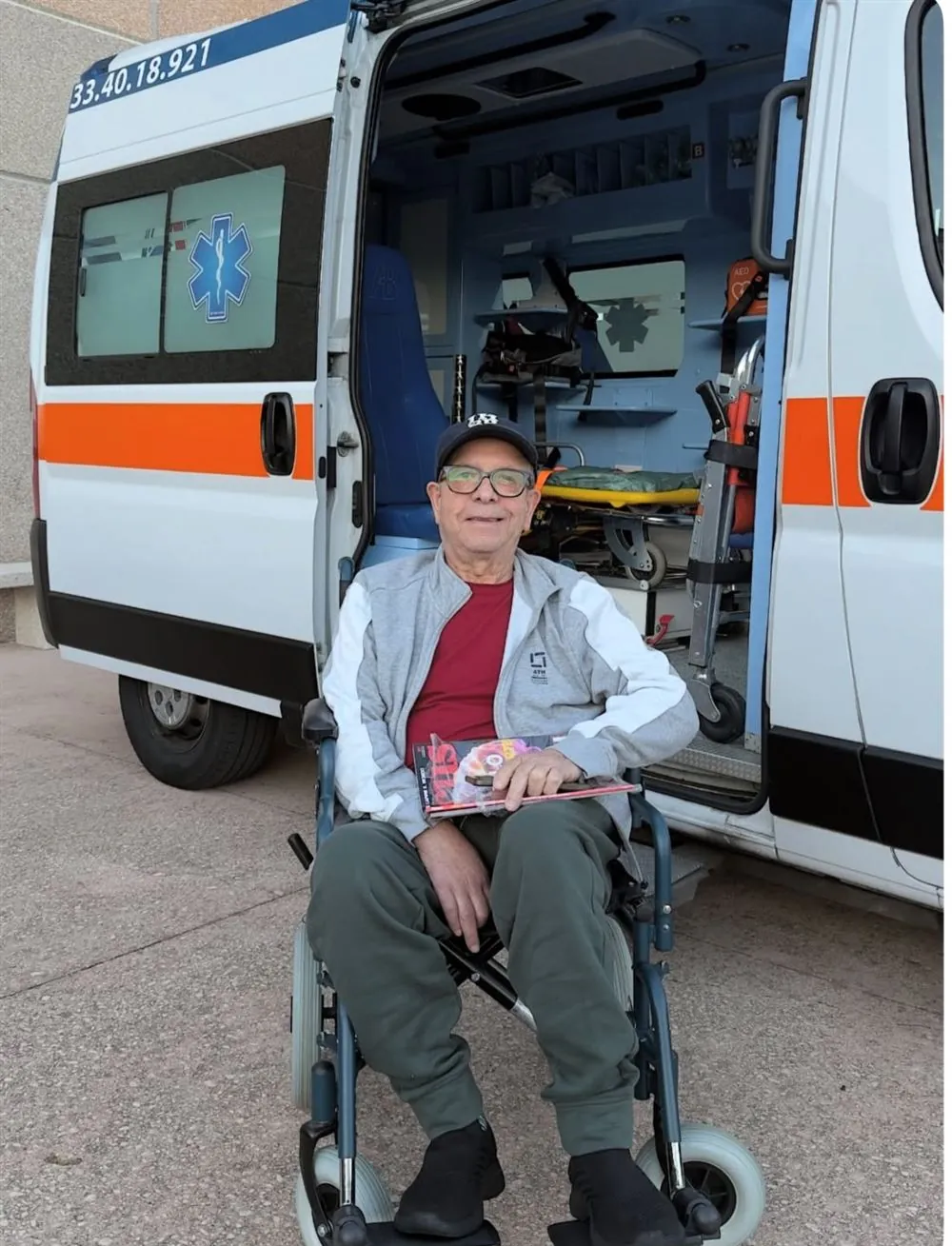 Un paziente sorridente sulla sedia da trasporto davanti all'ambulanza ALPHA aperta, con barella e defibrillatore a bordo