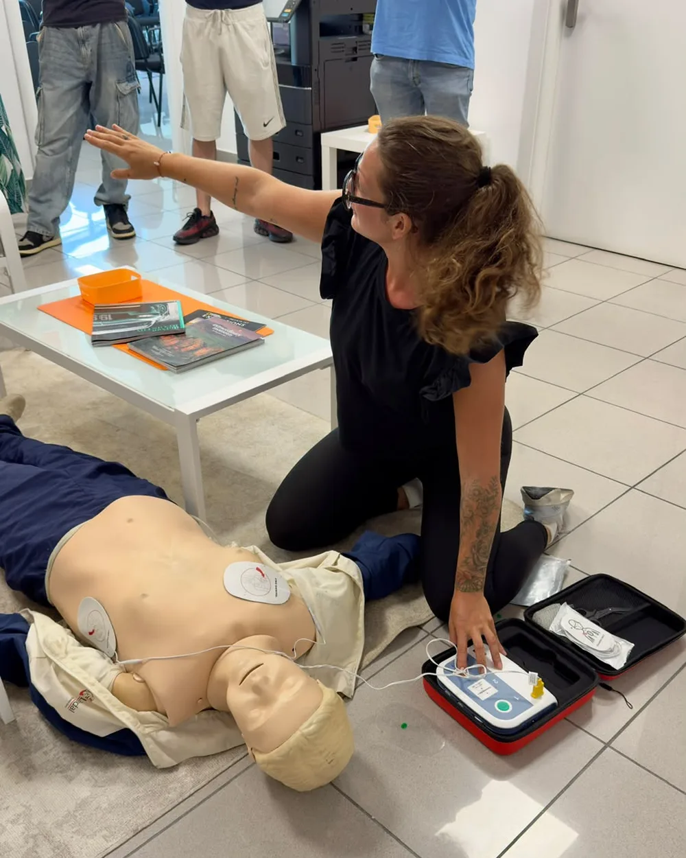 Un'istruttrice BasicLife inginocchiata accanto a un manichino spiega l'uso del defibrillatore didattico ai corsisti di un'azienda