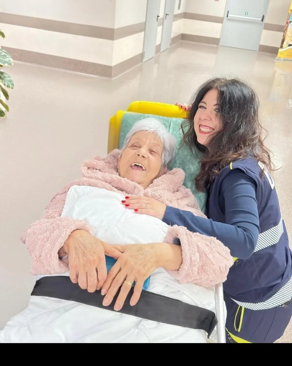 Un'operatrice ALPHA sorride abbracciata a una paziente anziana sulla barella in ospedale