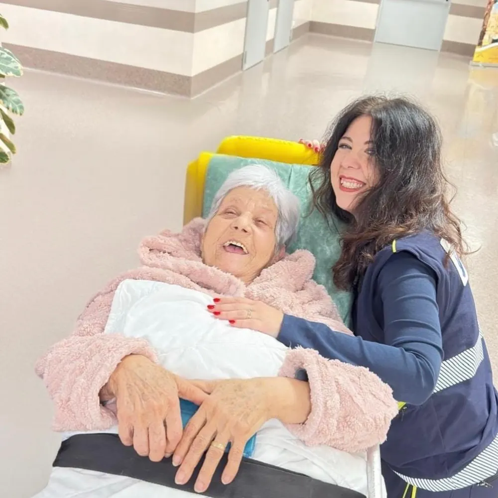 Operatrice ALPHA sorride accanto a una signora anziana sulla barella nel corridoio dell'ospedale