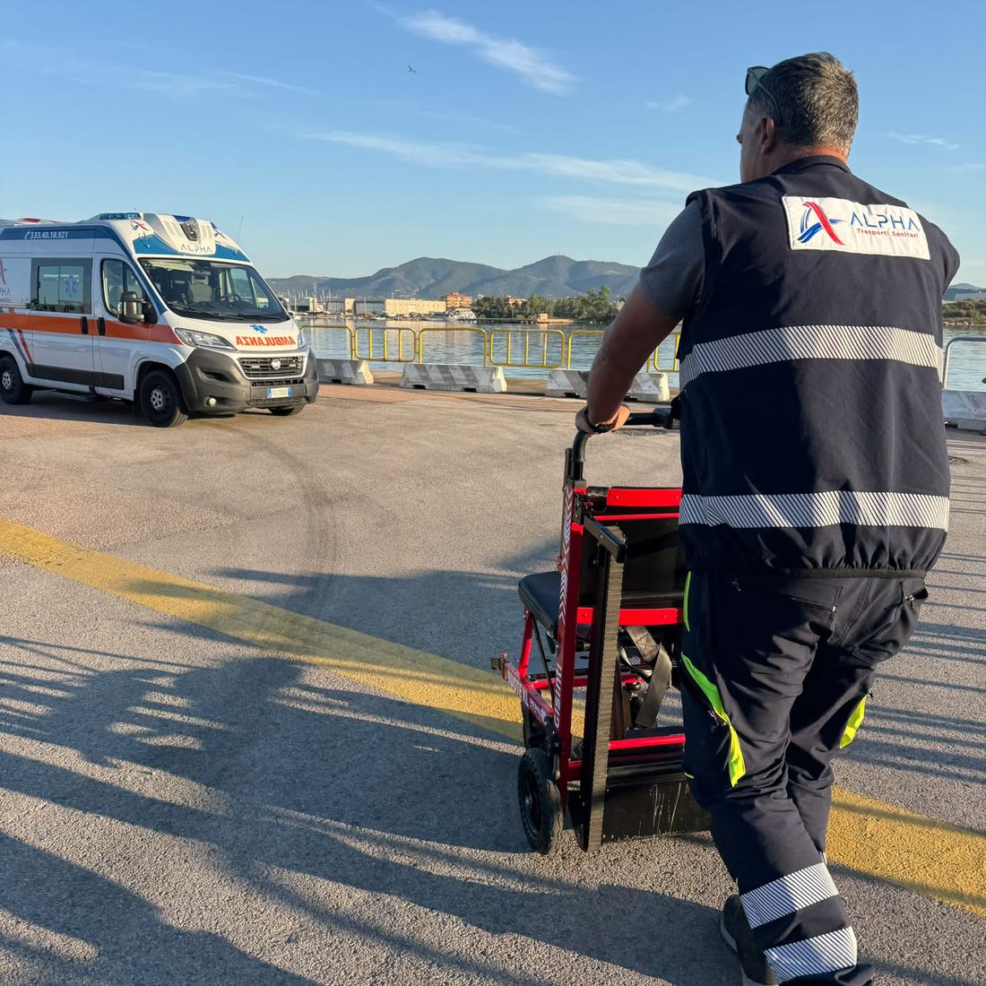 Un operatore ALPHA spinge la sedia saliscale elettrica a cingoli verso l'ambulanza, sulla banchina del porto