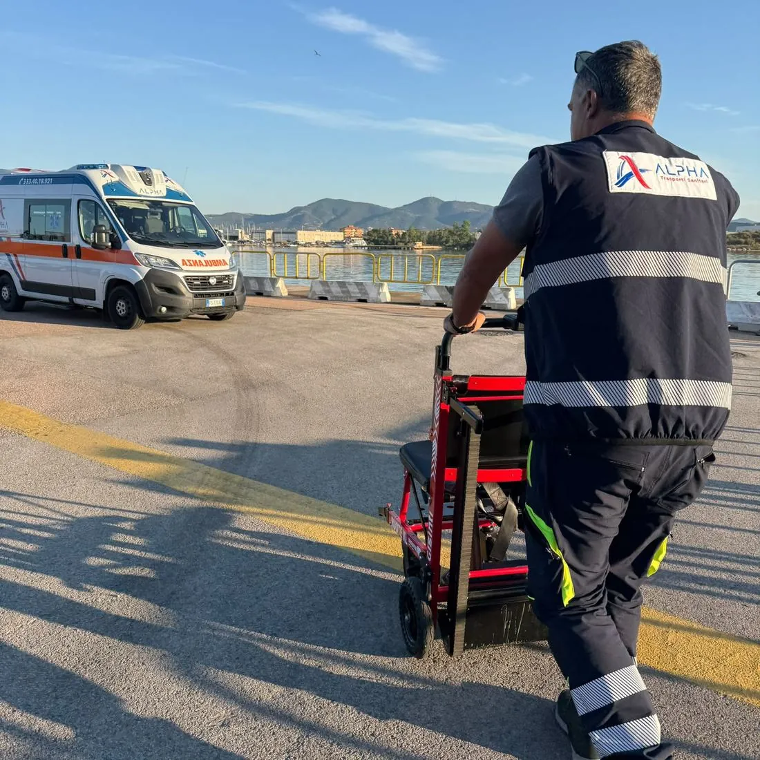 Un operatore ALPHA spinge la sedia saliscale elettrica a cingoli verso l'ambulanza, sulla banchina del porto